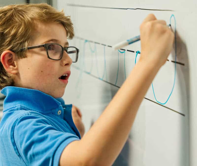 Young boy with glasses practicing handwriting on a whiteboard.